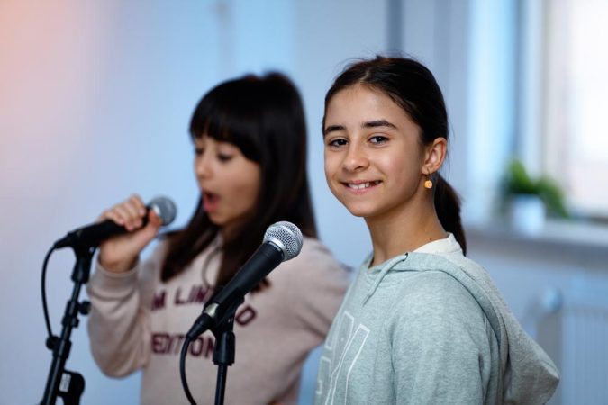 Kaksi iloista koululaistyttöä laulaa seisten mikrofoneihin
